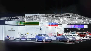 Auf einer hell beleuchteten Vienna Autoshow 2020 sind mehrere BMW Fahrzeuge auf Plattformen ausgestellt. Schlicht gehaltene Großprints, Markenzeichen und Informationsbanner sind im Hintergrund oder hängen über den BMW Modellen und Hybridfahrzeugen. Man sieht Menschen, die umhergehen und die Autos beobachten.
