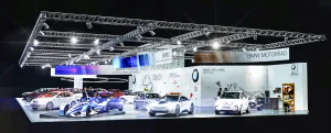 Ein hell erleuchteter BMW Messestand zeigt mehrere Autos und Motorräder, darunter einen blauen Formel-E-Rennwagen, unter einem modernen Metallrahmen auf der Vienna Autoshow 2020. Das BMW-Logo und die Beschilderung sind an den Standwänden zu sehen.