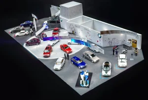 Vogelperspektive des BMW Messestandes auf der Vienna Autoshow 2020. Auf dem Messestand sind mehrere Fahrzeuge ausgestellt. Besucher stehen um die Autos herum, und moderne Beleuchtung und Großprints schmücken den Hintergrund. Im Vordergrund gehen Menschen auf einem roten Teppich.