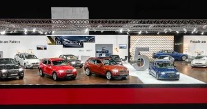 BMW Austria Messestand, Zentralbereich, auf der Vienna Autoshow 2017 mit verschiedenen BMW Fahrzeugen in unterschiedlichen Farben, darunter grün, lila, rot und blau. Im Zentrum ist die BMW 5er Insel mit einem großer Ziffer 5 aus Beton. Der Bereich ist gut beleuchtet, und es sind Branding- und Informationsschilder sichtbar.