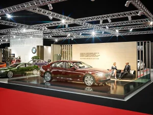 Auf der Messe Vienna Auto Show 2018 ist ein BMW 7 Modell unter hellen Lichtern ausgestellt, eine Frau und ein Mann sitzen auf einem Sofa und unterhalten sich.