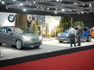 Zwei Männer stehen neben einem blauen BMW X3 auf Vienna Autoshow 2018. Ein weiterer grauer BMW X3 ist in der Nähe ausgestellt. Im Hintergrund sind Stadtansichten zu sehen und das BMW-Logo ist an der Wand zu erkennen.