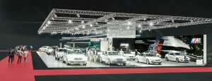 Vogelperspektive des BMW Messestandes auf der Vienna Autoshow 2019 ausgestellt. Auf dem Messestand sind mehrere Fahrzeuge ausgestellt. Besucher stehen um die Autos herum, und moderne Beleuchtung und Großprints schmücken den Hintergrund. Im Vordergrund gehen Menschen auf einem roten Teppich.