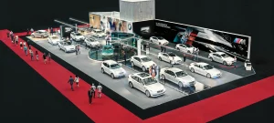 Vogelperspektive des BMW Messestandes auf der Vienna Autoshow 2019 ausgestellt. Auf dem Messestand sind mehrere Fahrzeuge ausgestellt. Besucher stehen um die Autos herum, und moderne Beleuchtung und Großprints schmücken den Hintergrund. Im Vordergrund gehen Menschen auf einem roten Teppich.
