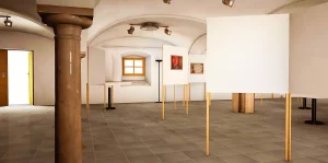 Visualisierun einer Kunstausstellung im Säulenfoyer des Schauspielhauses in Salzburg. Im Raum mit Säulen stehen Wände und Tische auf denen die Kunstwerke präsentiert werden. Auf den Wänden hängen Bilder.