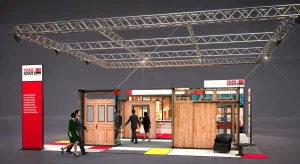 Messestand mit Holztüren und einem Pult und Boden im Mondrian Design. Im Hintergrund ist eine Lounge mit dem Großprint einer Bar. Auf dem Messestand gehen Menschen herum. Links ist ein Pylon mit dem Feuerschutz Team Logo und Angebot