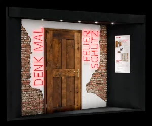 Messe Display von Feuerschutz team im einer Tür aus Holz in einem alten Mauerwerk aus Ziegeln. Lins und rechts der Tür ist eine Schrift aus roten Neonröhren "Denk mal" und "Feuer Schutz". Rechts ist ein Plakat mir Text und Bildern.