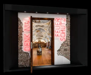 Messe Display von Feuerschutz team im einer Tür aus Holz in einem alten Mauerwerk aus Ziegeln. Die Tür ist offen und zeigt in eine alte Bibliothek. Lins und rechts der Tür ist eine Schrift aus roten Neonröhren "Denk mal" und "Feuer Schutz".