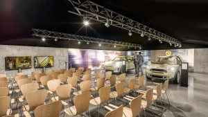 In einem Präsentations- und Schulungsraum stehen Stühle für die Teilnehmer vor zwei LED Bildschirmen. Im Hintergrund sind zwei MINI Electric Fahrzeuge. Die Wände sind mit Großprints gestaltet. Zwischen den MINIs stehen zwei Männer in Businessanzügen