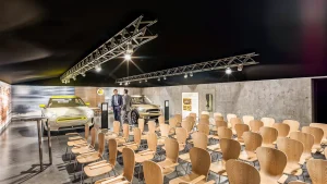 In einem Präsentations- und Schulungsraum stehen Stühle für die Teilnehmer. Im Hintergrund sind zwei MINI Electric Fahrzeuge und eine Ladestation. Die Wände sind mit Großprints gestaltet, zwischen den MINIs stehen zwei Männer in Businessanzügen.
