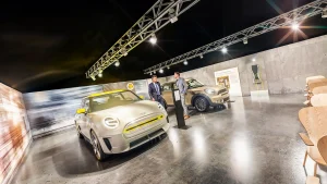 In einem Präsentations- und Schulungsraum stehen zwei MINI Electric Fahrzeuge und eine Ladestation. Die Wände sind mit Großprints gestaltet. Zwischen den MINI Fahrzeugen stehen zwei Männer in Businessanzügen.