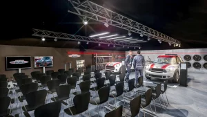 In einem Präsentations- und Schulungsraum stehen zwei MINI John Cooper Works Fahrzeuge. Die Wände sind mit Teilen und Felgen geschmückt. Vor den MINI Fahrzeugen stehen zwei Männer in Businessanzügen. Im Vordergrund stehen schwarze Stühle vor zwei LED Bildschirmen für die Teilnehmer.