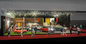 Menschen gehen an dem MINI Messestand auf der Vienna Autoshow vorbei, bei der verschiedene MINI-Modelle präsentiert werden. Die Ausstellung findet in einer Halle statt, die mit heller Beleuchtung und einem roten Teppich ausgestattet ist.