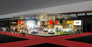 Menschen gehen an einer Autoausstellung vorbei, bei der verschiedene MINI-Modelle präsentiert werden. Die Ausstellung findet in einer Halle statt, die mit heller Beleuchtung und einem roten Teppich ausgestattet ist.