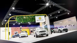 Auf der Vienna Autoshow 2020 sind mehrere MINI Fahrzeuge unter einem Hängegarten mit dem Display "MINI Electric" rechts oben. Ein MINI John Cooper Works steht vor einer Betonwand mit Bildern aus der MINI Geschichte.