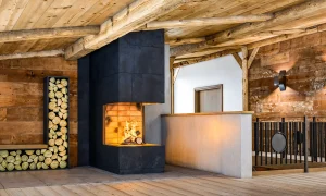 Ein Blockhausinterieur mit Kamin, in dem ein Feuer brennt. Links ist eine Bank mit gestapeltem Brennholz. Rechts ist eine Geländer aus Schwarstahl. Der Kamin ist ebenfalls mit Schwarzstahl verkleidet.