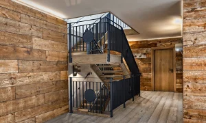 Eine Treppe in einem Blockhaus. Wände sind mit gekalktem Altholz verkleidet. In der Mitte ist eine Stiege mit einem Geländer aus geöltem Schwarzstahl.