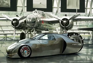 Futuristisches Elektroauto mit drei Rädern. Ein großes Antriebsrad am Heck. Karosserie ist aus Carbon. Auto steht im Hangar 7 in Salzburg vor einem B52 WWII Bomber. Seitenansicht. Das Hintergrundfoto wurde von Carsten Müller erstellt.