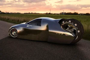 Futuristisches Elektroauto mit drei Rädern. Ein großes Antriebsrad am Heck. Karosserie ist aus Carbon. Auto steht auf einer Straße vor einer Wiese beim Sonnenuntergag. Im Hintergrund sind Windräder zu sehen.